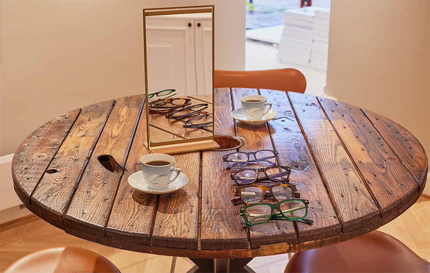 Hyggeligt træord med briller der ligger på række og en mågestel kop med god kaffe i. God tid til at prøve briller.